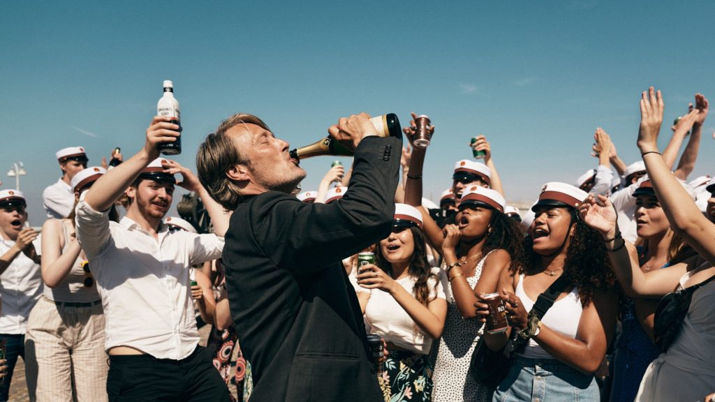 Still from the film Another Round, profile of a man drinking from a champagne bottle with a crowd of young people behind him