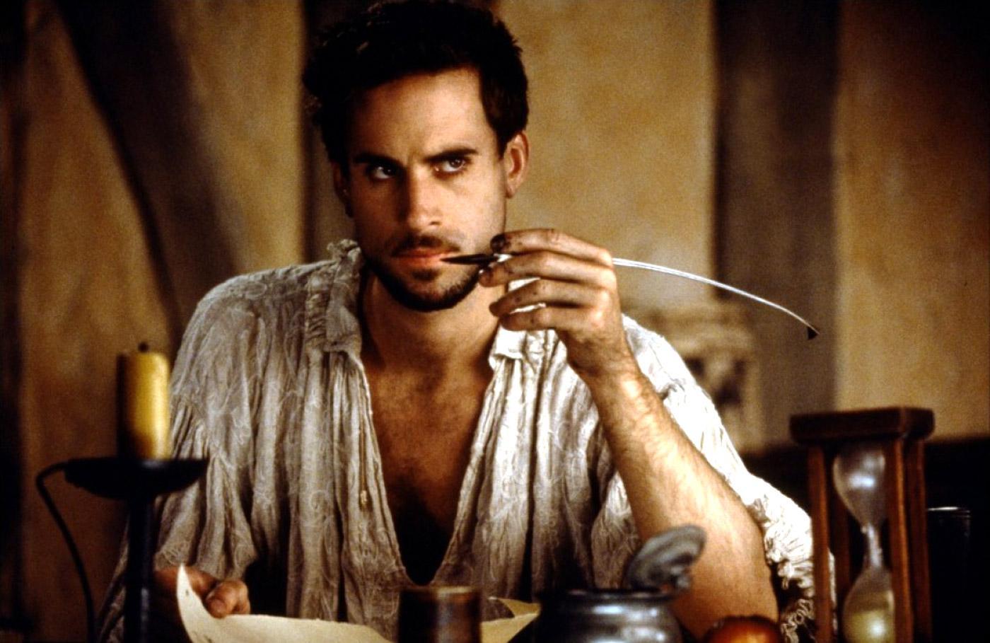 Still from Shakespeare in Love, mid shot of a Renaissance man sitting at a desk with a quill