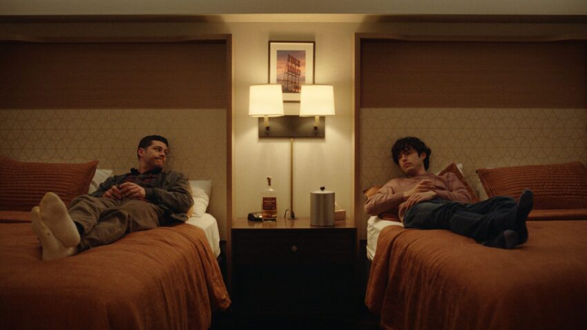 Still from Twinless (2025). Medium shot of two men sitting up in side by side hotel beds, looking at each other