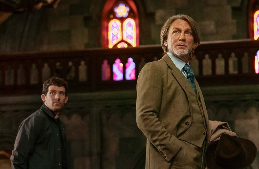 Still from Wake Up Dead Man (2025). Man in a brown, windowpane checked three piece suit in the foreground, a younger man in a black priest's cassock behind him to the left